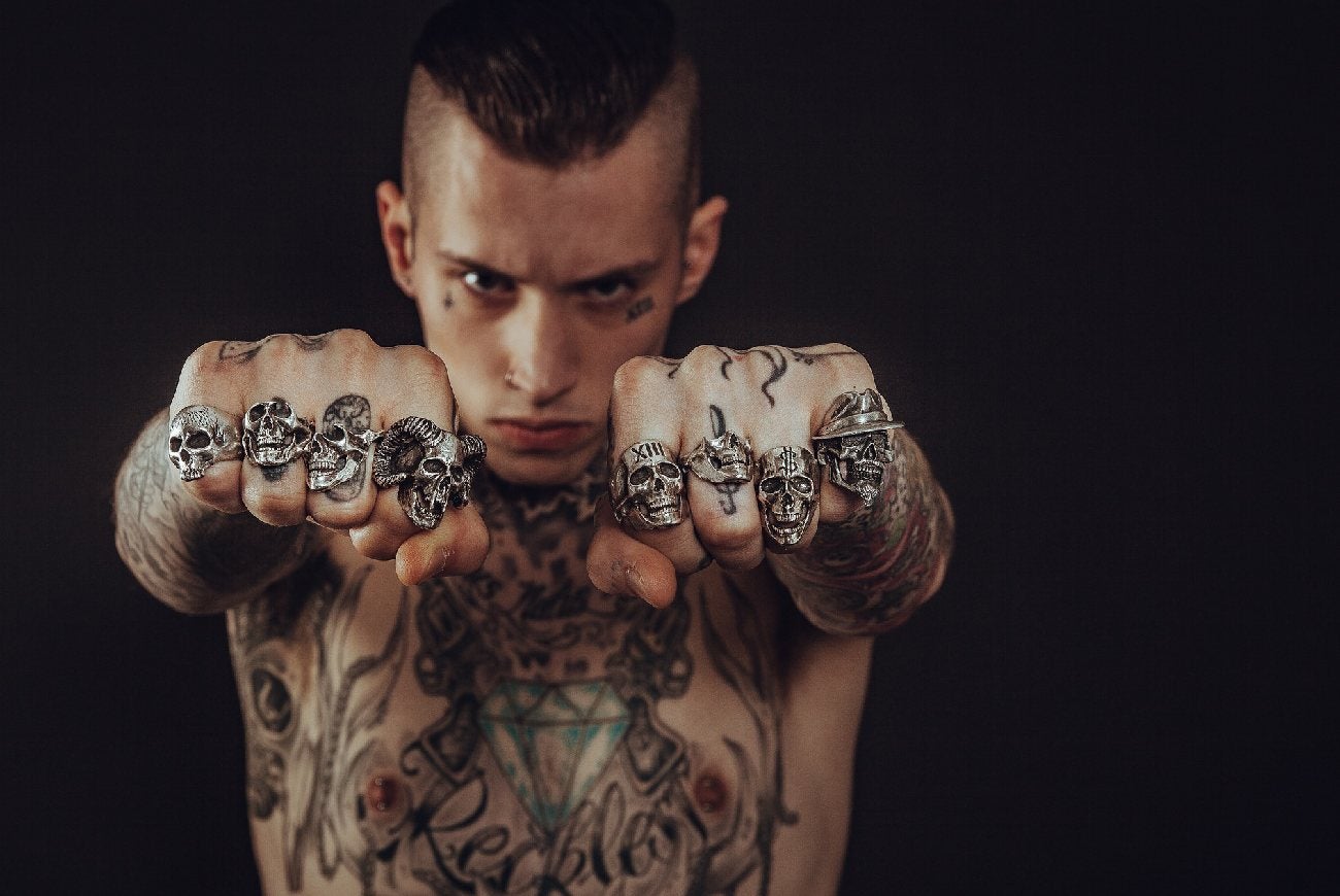 Hombre con los puños extendidos y el cuerpo lleno de tatuajes y anillos en sus manos