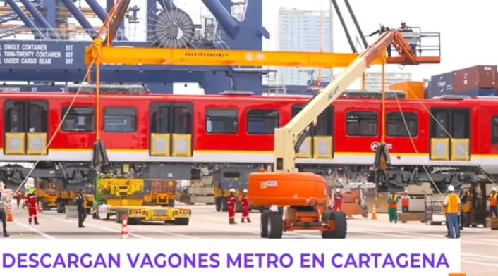 Vista lateral del tren mientras es bajado cuidadosamente del buque. Varias personas del equipo técnico coordinan la maniobra en tierra, con contenedores marítimos de fondo. Vista lateral del tren mientras es bajado cuidadosamente del buque. Varias personas del equipo técnico coordinan la maniobra en tierra, con contenedores marítimos de fondo.